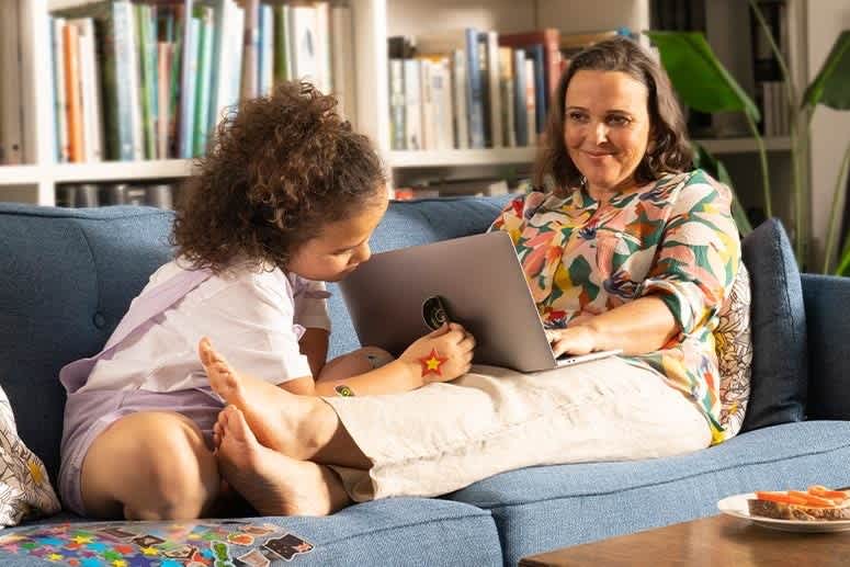 a girl applying a sticker to her mother's laptop as her mother watches on smiling