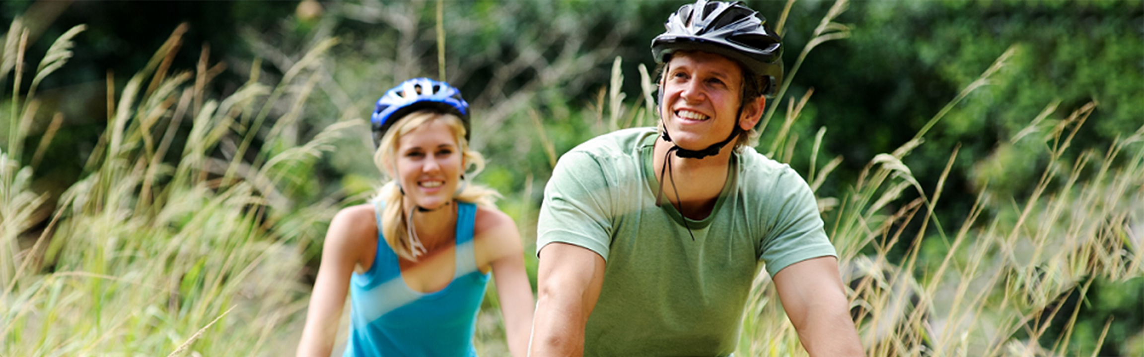 A man and a woman smile gleefully as they ride their bicycles in a field. 